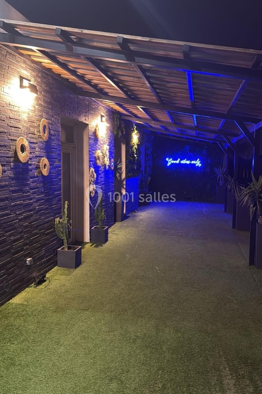 Allée couverte avec sol en gazon artificiel, murs en pierre décorés et enseigne lumineuse bleue au fond.