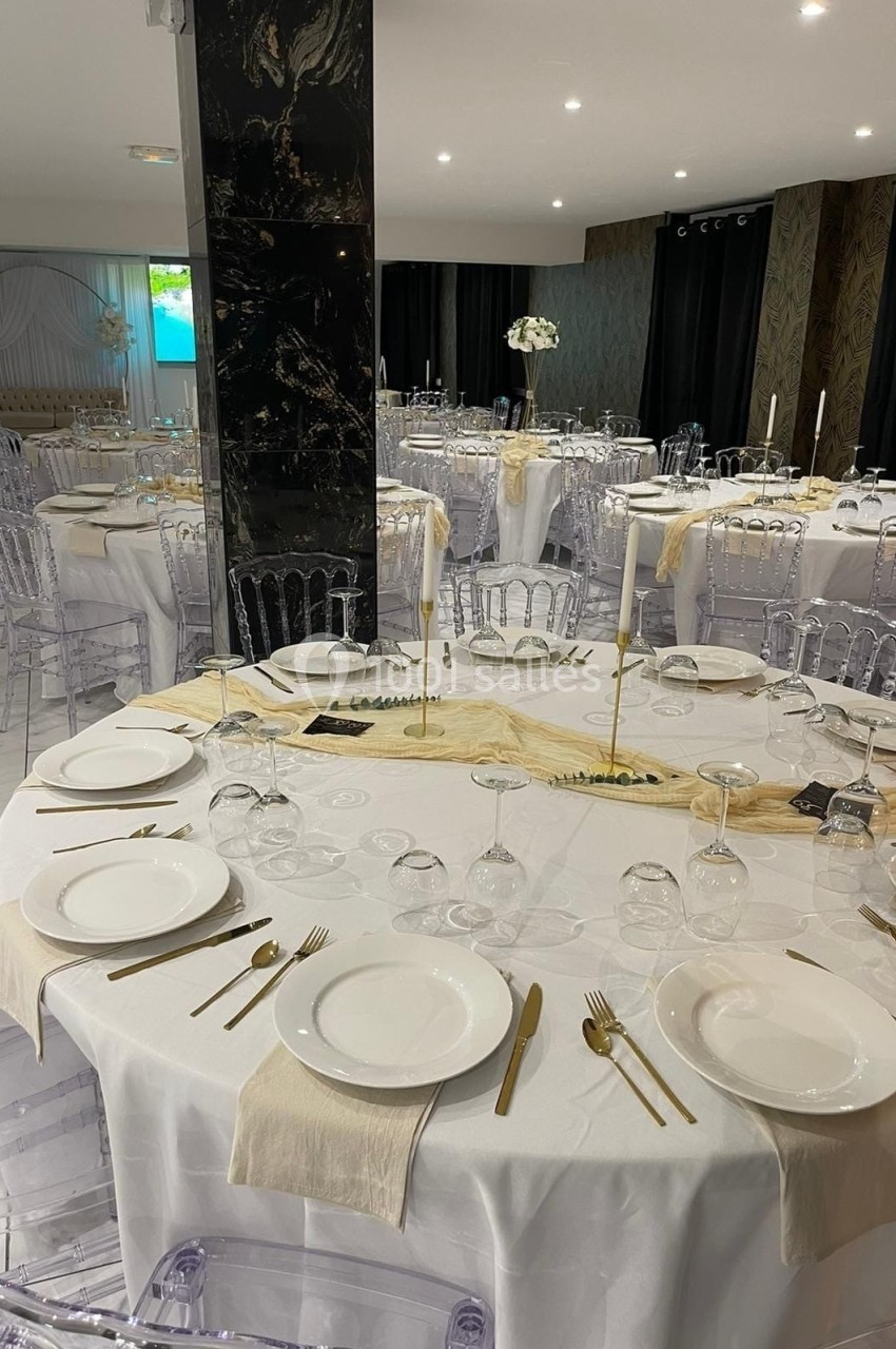 Salle de réception avec tables rondes dressées, nappes blanches, couverts dorés et chaises transparentes.