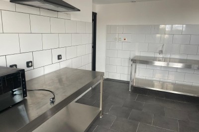 Cuisine professionnelle avec plan de travail en inox, carrelage mural blanc, sol en carrelage gris et équipements basiques.
