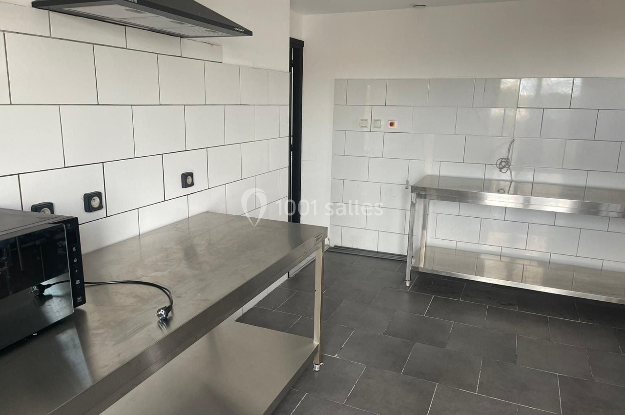 Cuisine professionnelle avec plan de travail en inox, carrelage mural blanc, sol en carrelage gris et équipements basiques.