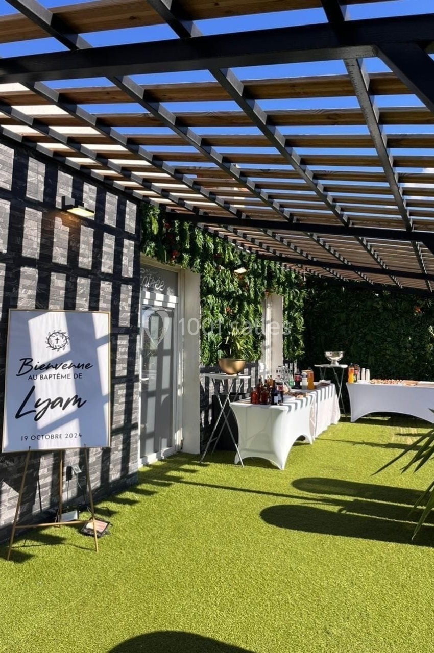 Terrasse décorée avec pergola, panneau de bienvenue pour un baptême, tables avec boissons et buffet, mur végétal en arrière…