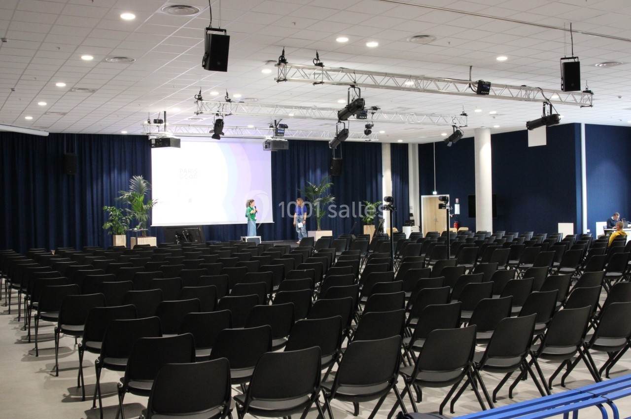 Salle de conférence vide avec des rangées de chaises noires, un écran de projection allumé et quelques personnes sur scène.