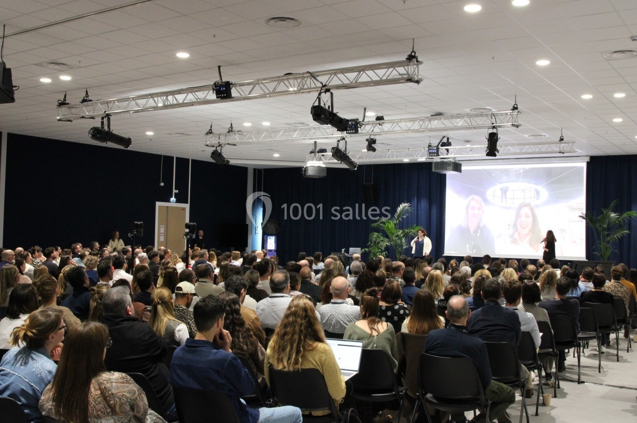 Salle de conférence remplie, avec des intervenants s'exprimant devant un public attentif et un écran projeté.