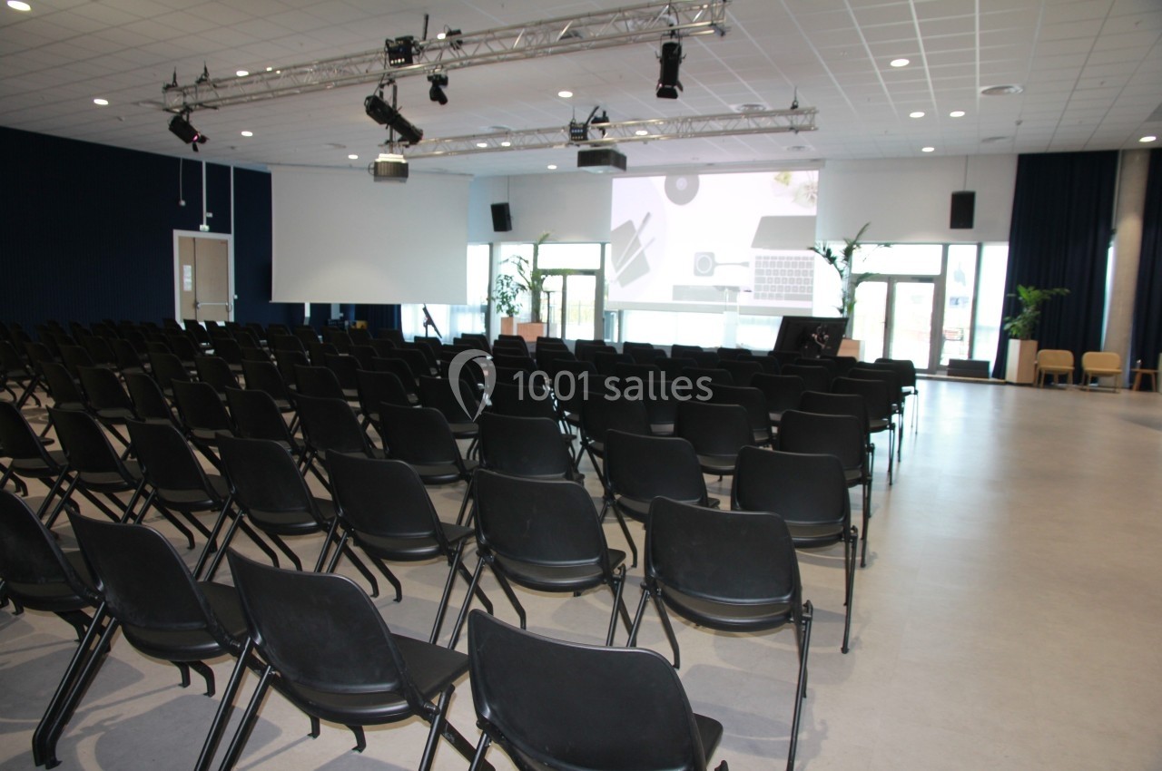 Salle de conférence vide avec des rangées de chaises noires, un écran de projection et des équipements audiovisuels.