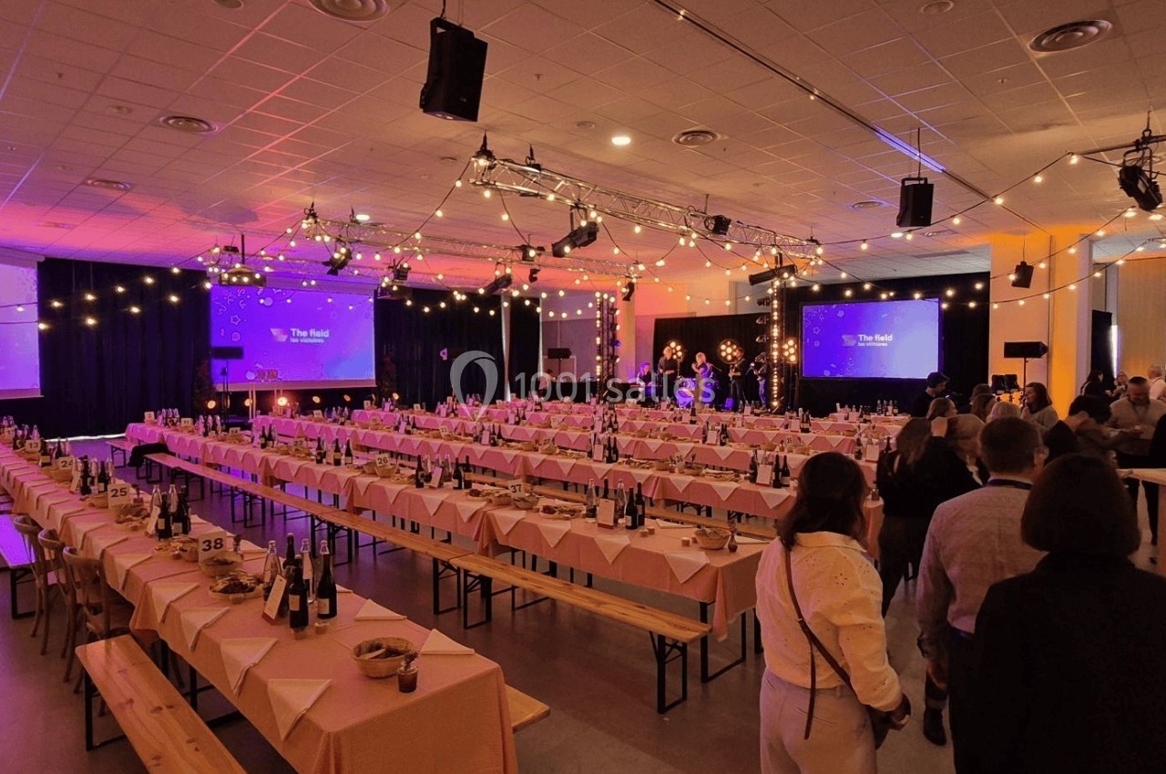 Salle de réception décorée avec des guirlandes lumineuses, tables dressées et scène en arrière-plan.
