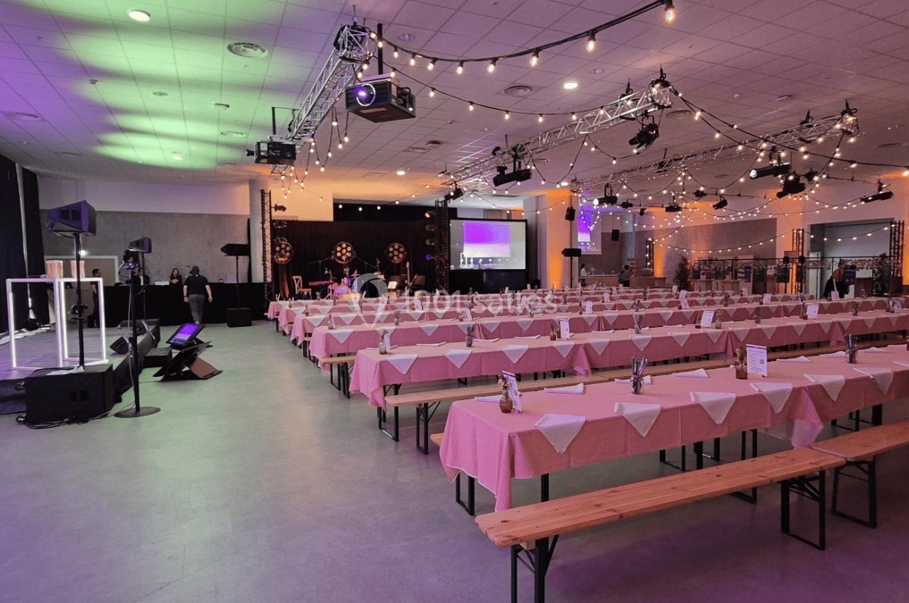 Salle de réception décorée avec des guirlandes lumineuses, tables alignées avec nappes roses et scène équipée au fond.