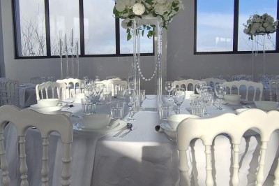 Miniature Location salle Épinay-sur-Seine (Seine-Saint-Denis) - Salle Delena #3 Salle lumineuse avec table ronde dressée, chaises blanches et centre de table floral sous de grandes fenêtres.