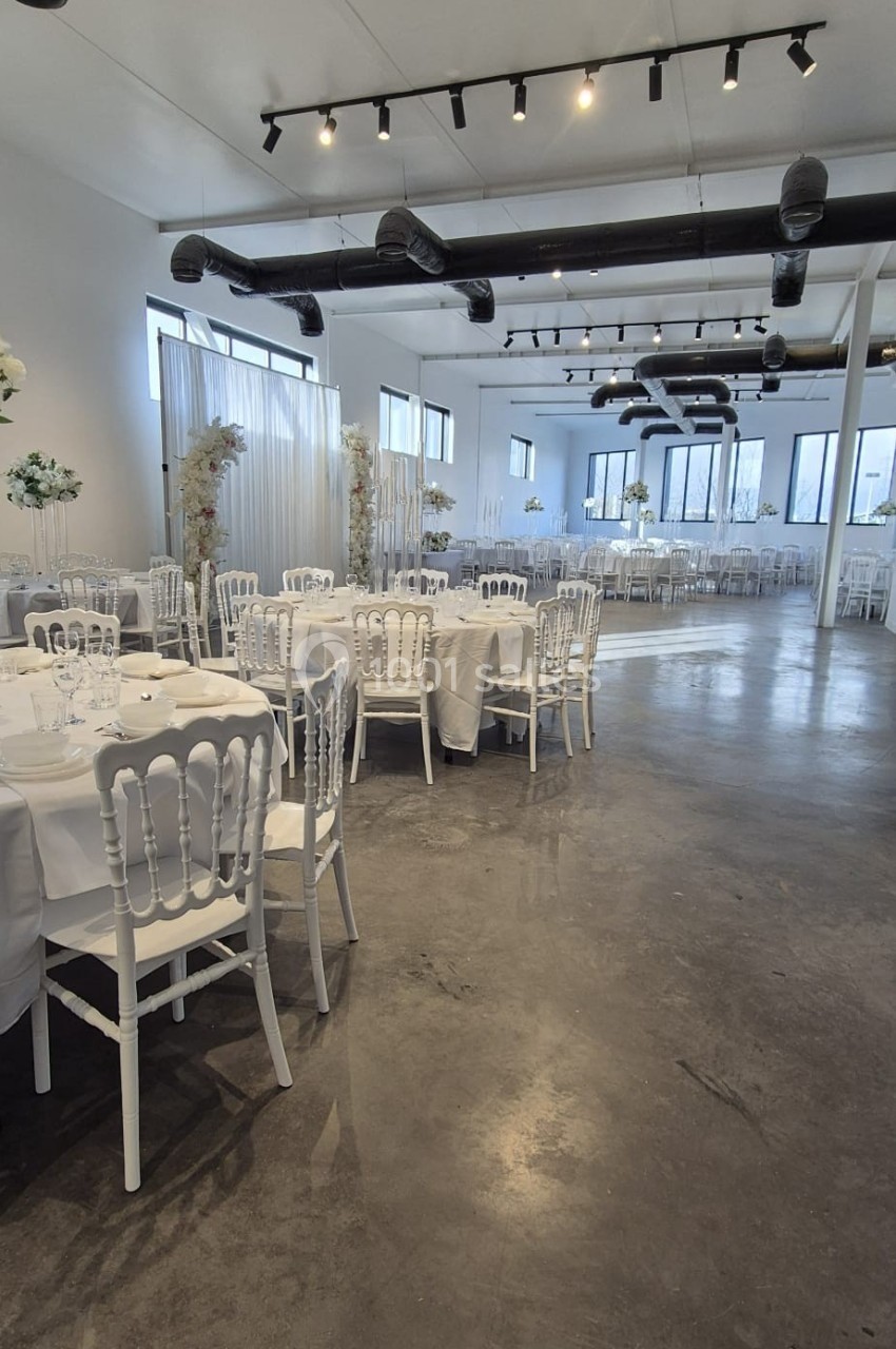 Salle de réception lumineuse avec tables rondes dressées, chaises blanches et décorations florales.