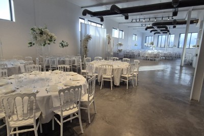 Miniature Location salle Épinay-sur-Seine (Seine-Saint-Denis) - Salle Delena #4 Salle lumineuse avec table ronde dressée, chaises blanches et centre de table floral sous de grandes fenêtres.