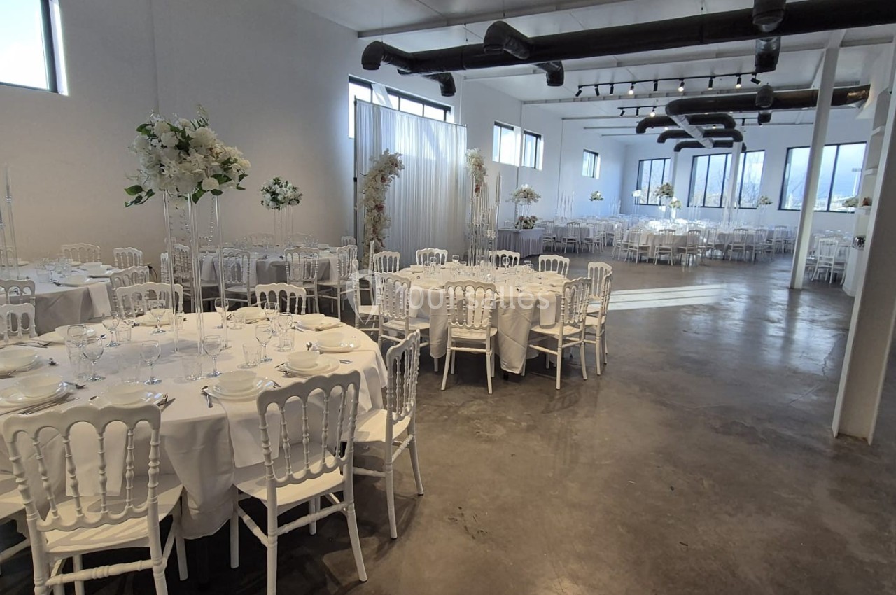 Salle de réception lumineuse avec tables rondes dressées, chaises blanches et décorations florales élégantes.