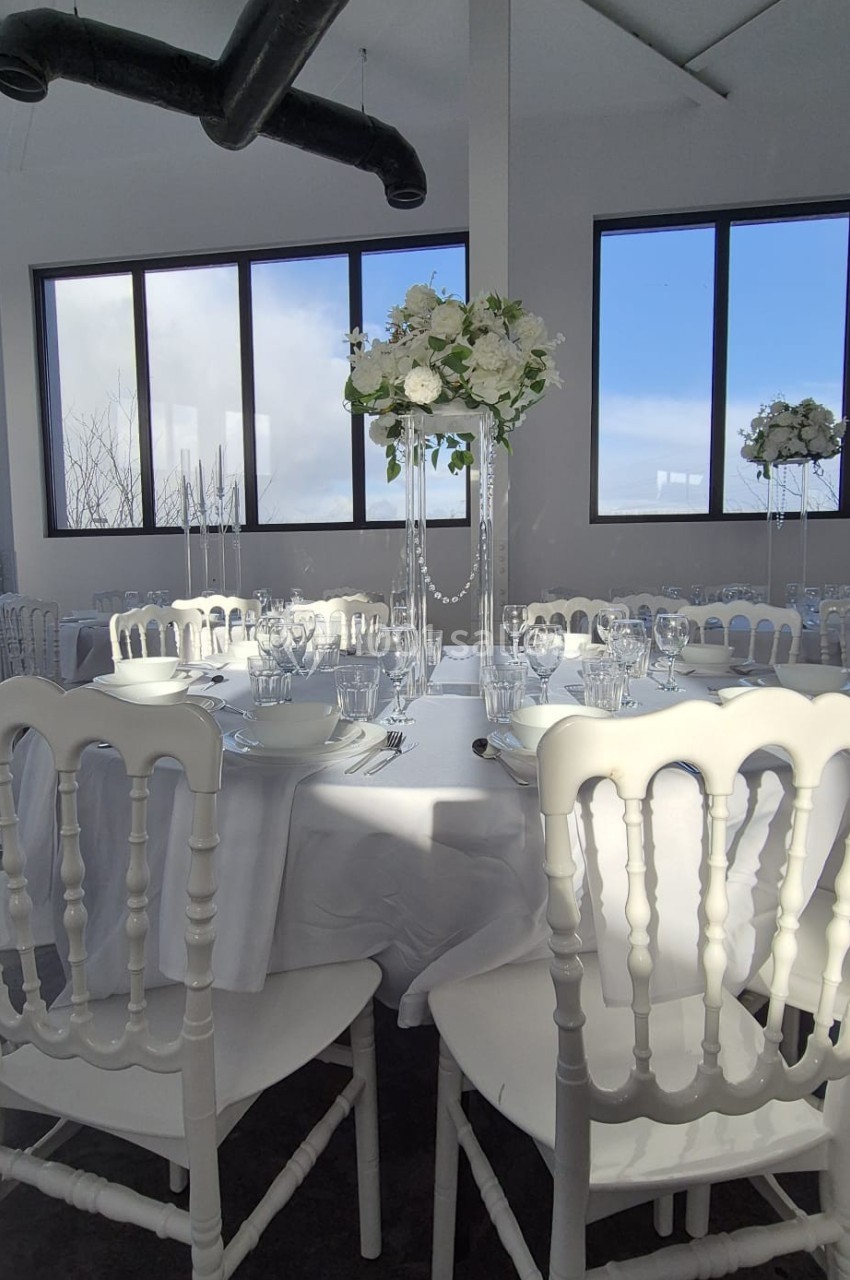 Salle lumineuse avec table ronde dressée, nappée de blanc, décorée de fleurs et entourée de chaises blanches.