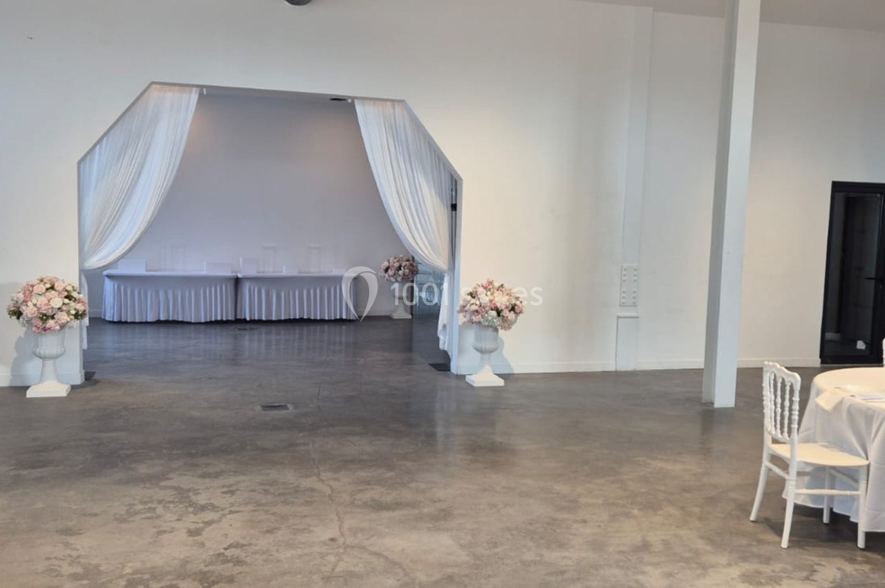 Salle de réception lumineuse avec sol en béton, rideaux blancs encadrant une ouverture et tables décorées de fleurs.