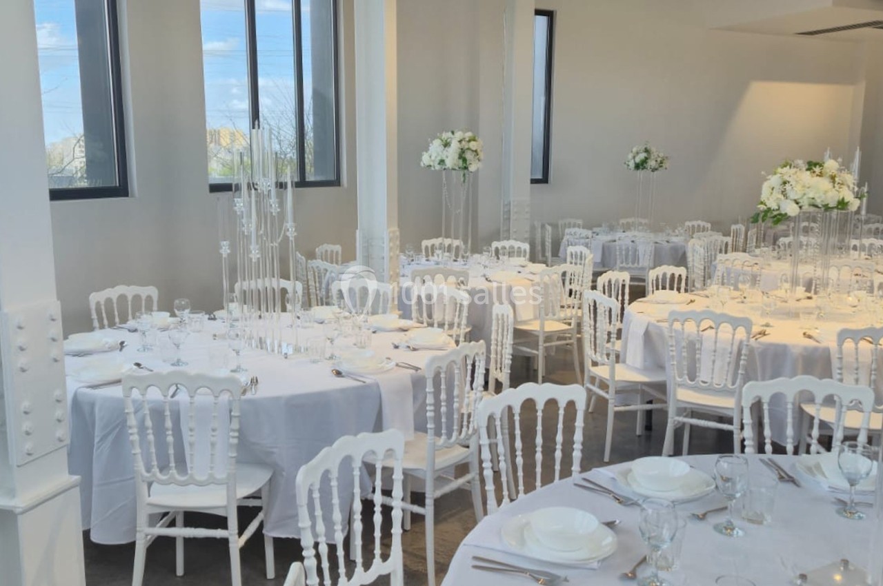 Salle de réception lumineuse avec tables rondes dressées, nappes blanches et décorations florales élégantes.