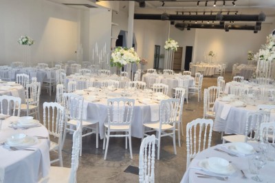 Miniature Location salle Épinay-sur-Seine (Seine-Saint-Denis) - Salle Delena #9 Salle lumineuse avec table ronde dressée, chaises blanches et centre de table floral sous de grandes fenêtres.