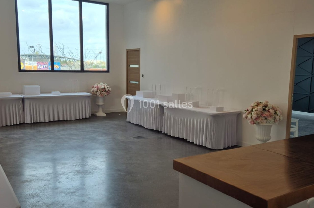 Salle lumineuse avec grandes fenêtres, tables drapées de blanc, décorations florales et sol en béton poli.