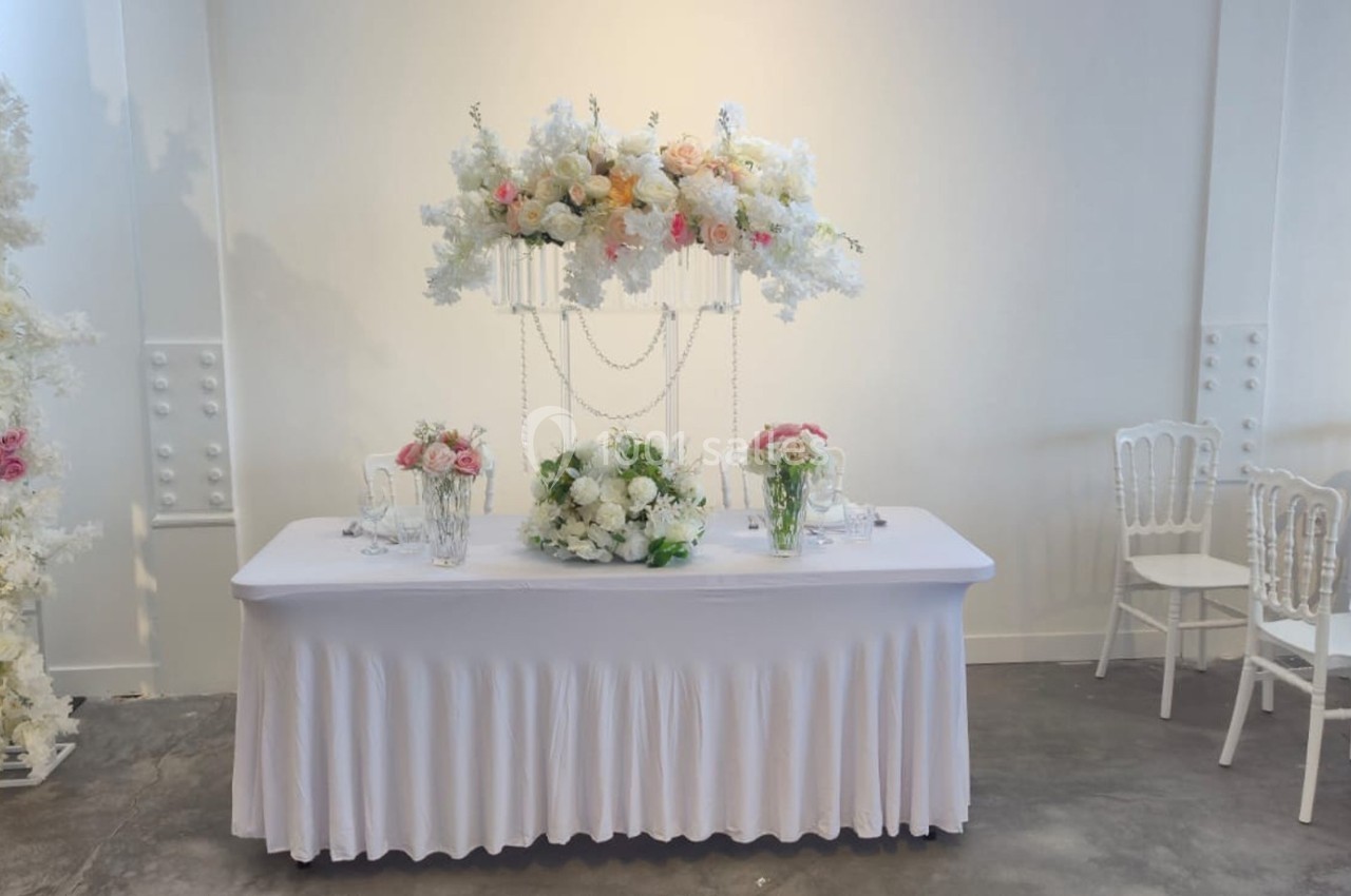 Table décorée pour un événement avec nappes blanches, fleurs blanches et roses, et chaises alignées en arrière-plan.