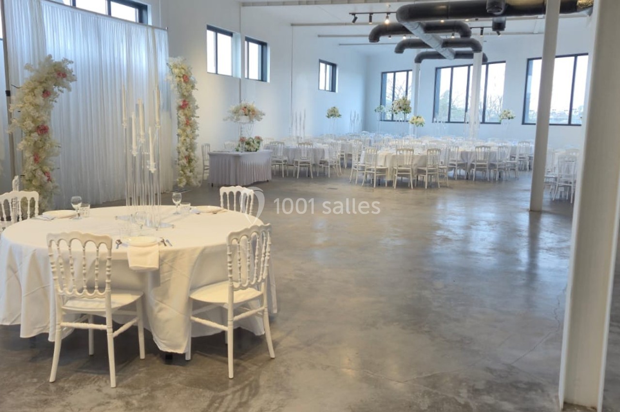 Salle de réception lumineuse avec tables rondes dressées, chaises blanches et décorations florales élégantes.