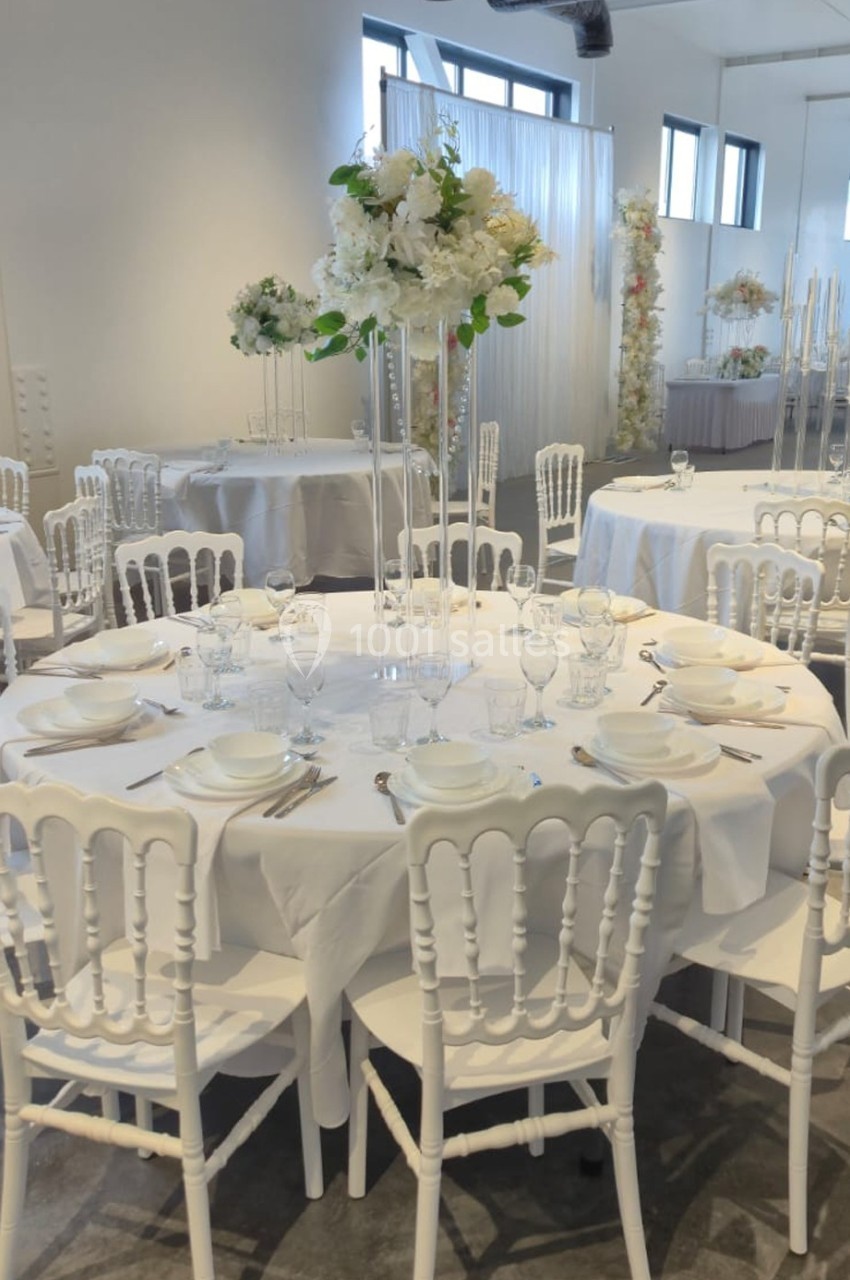 Salle de réception décorée avec des tables rondes, nappes blanches, chaises assorties et centres de table fleuris.