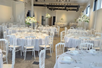 Miniature Location salle Épinay-sur-Seine (Seine-Saint-Denis) - Salle Delena #15 Salle lumineuse avec table ronde dressée, chaises blanches et centre de table floral sous de grandes fenêtres.