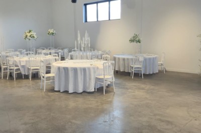 Miniature Location salle Épinay-sur-Seine (Seine-Saint-Denis) - Salle Delena #16 Salle lumineuse avec table ronde dressée, chaises blanches et centre de table floral sous de grandes fenêtres.