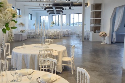 Miniature Location salle Épinay-sur-Seine (Seine-Saint-Denis) - Salle Delena #17 Salle lumineuse avec table ronde dressée, chaises blanches et centre de table floral sous de grandes fenêtres.