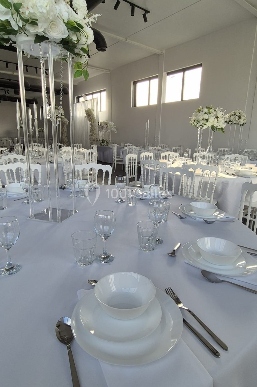 Salle de réception décorée avec des tables dressées, nappes blanches, vaisselle élégante et centres de table fleuris.