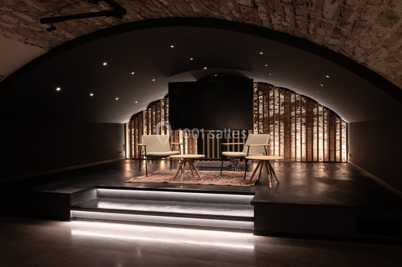 Salle voûtée avec éclairage tamisé, deux fauteuils et une table basse sur une estrade devant un mur en bois rétroéclairé.