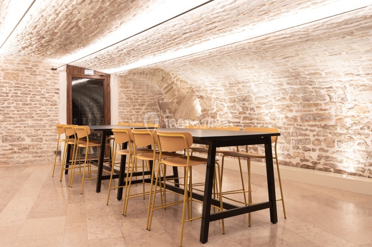 Salle voûtée en pierre avec une grande table noire entourée de chaises jaunes, éclairée par des lumières au plafond.
