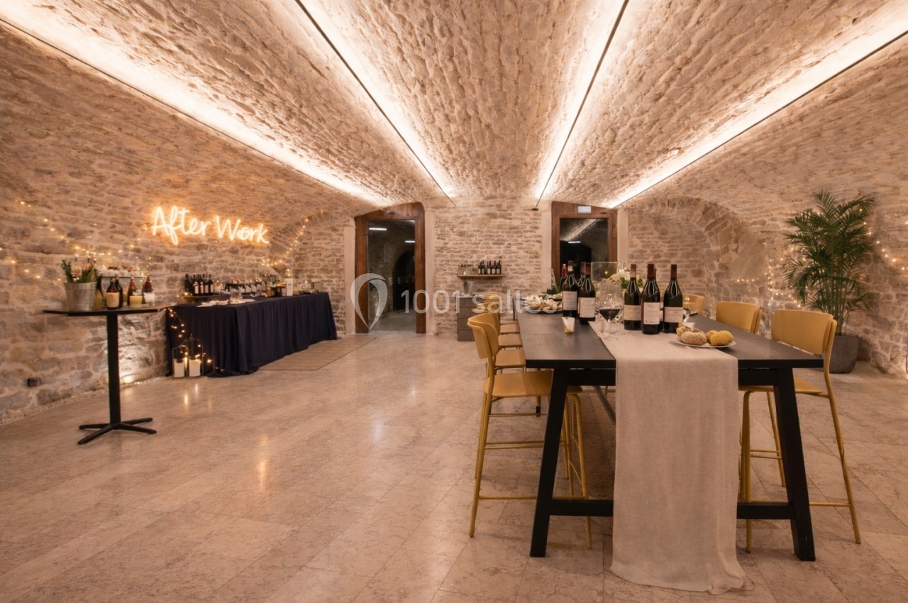 Salle voûtée en pierre aménagée pour un événement, avec tables décorées, bouteilles de vin et éclairage chaleureux.