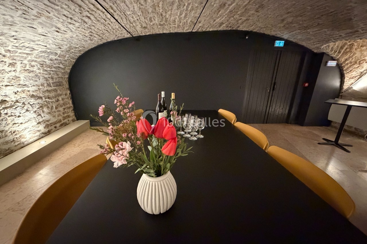 Table noire dans une cave voûtée, décorée d'un vase de fleurs, avec des verres et des bouteilles alignés.