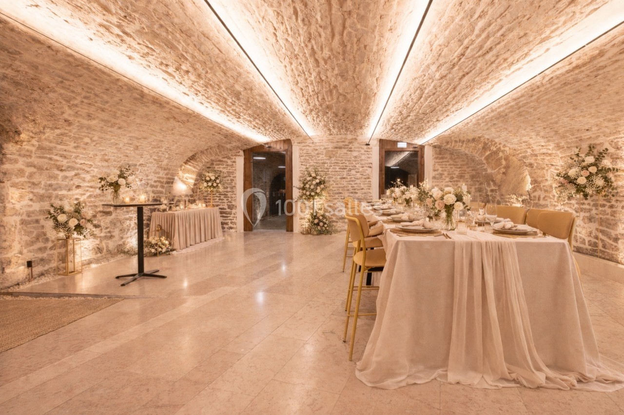Salle voûtée en pierre avec tables élégamment dressées, décorée de fleurs blanches et éclairée par des lumières chaleureuses.