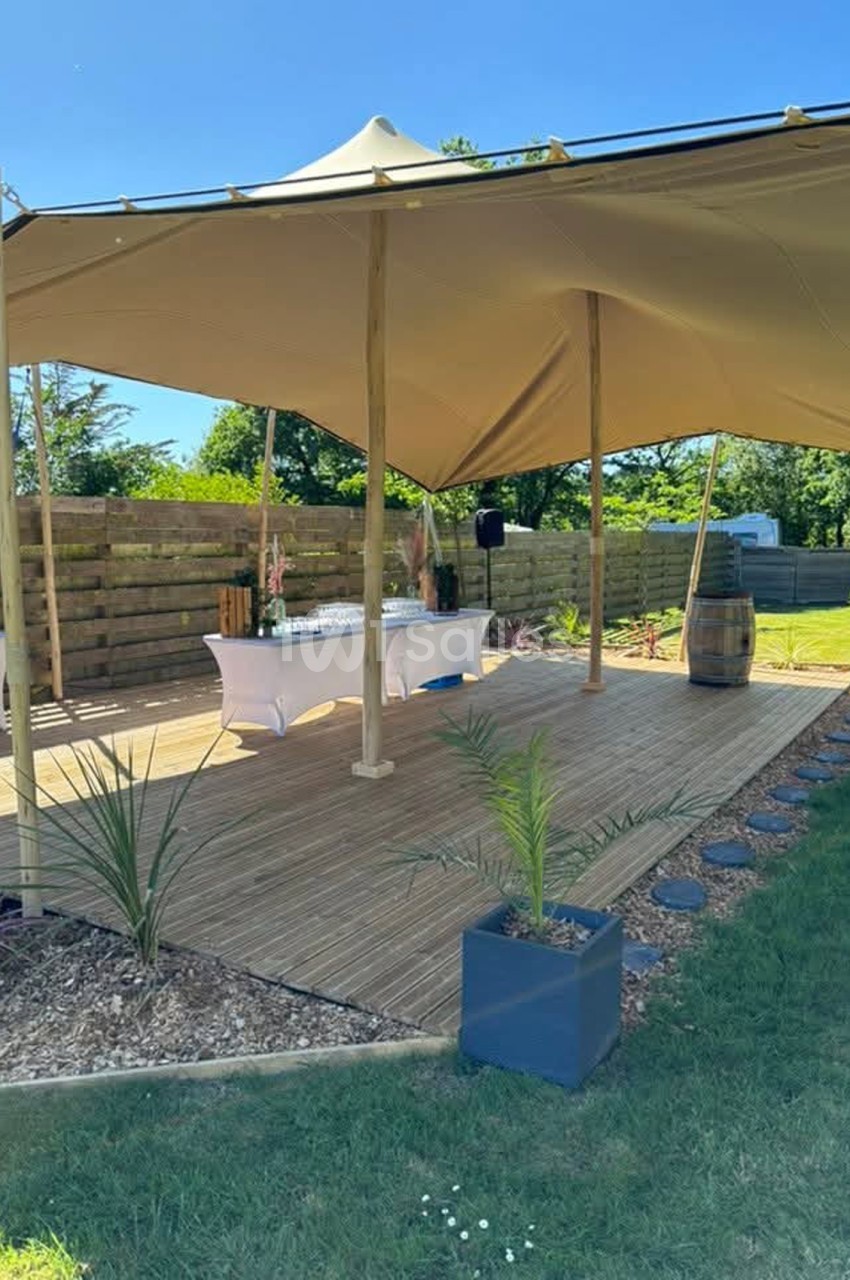 Terrasse en bois sous une grande voile tendue beige, avec deux tables nappées de blanc et une jardinière dans un extérieur arboré.