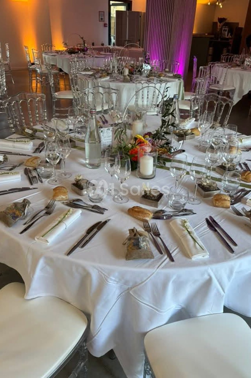 Table ronde nappée de blanc dressée pour un repas, avec chaises transparentes, verres, petits pains, bouquets de fleurs et bougies, dans une salle aux murs ocre éclairés. Let me check length: roughly 165 characters, too long. Need ≤125. Table ronde
