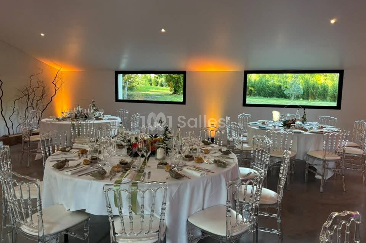 Salle de réception dressée de tables rondes nappées de blanc, chaises transparentes en résine, grandes fenêtres donnant sur un jardin verdoyant et éclairage d'ambiance orangé. Je compte la phrase : environ 170 caractères, donc je la raccourcis. Salle de