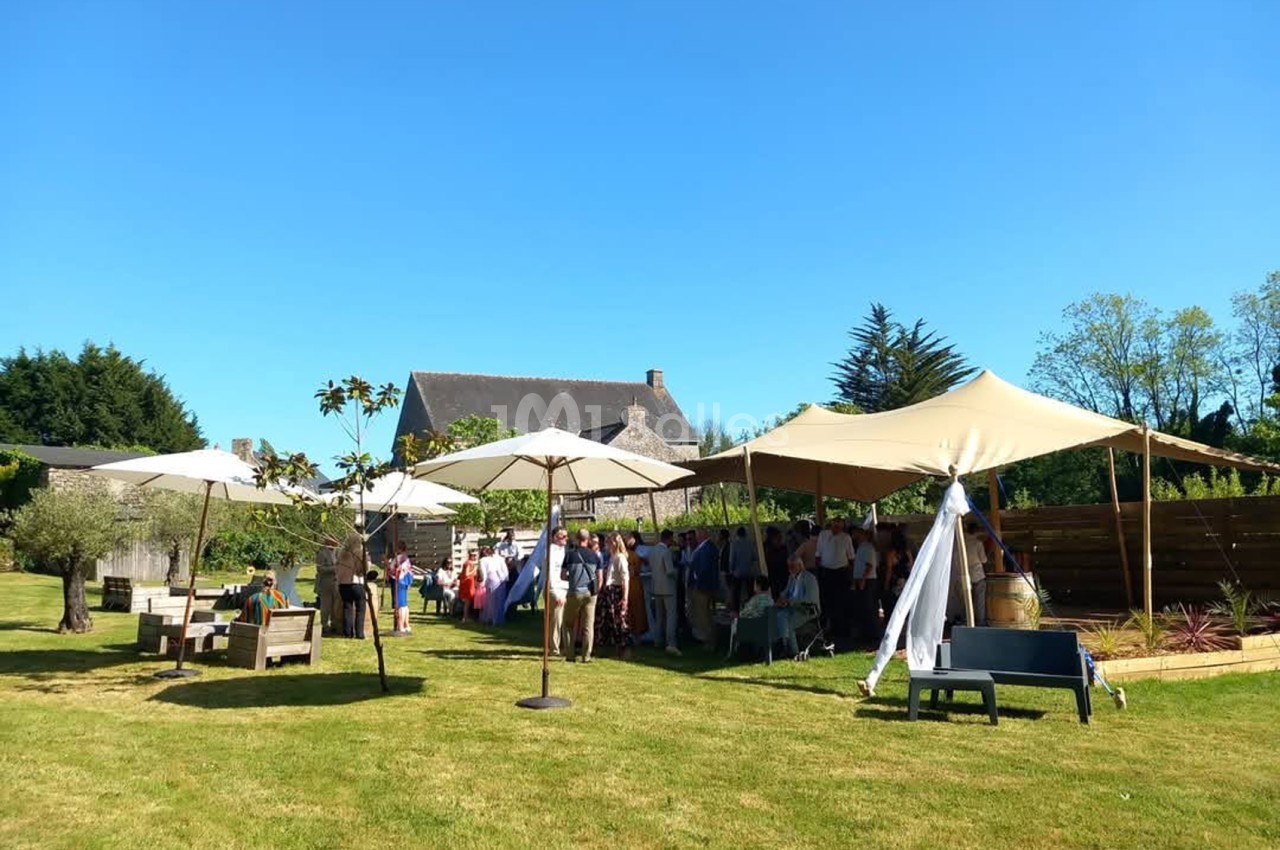 Jardin ensoleillé où des invités se tiennent sous une grande voile beige tendue, avec parasols et mobilier de jardin en bois sur la pelouse.