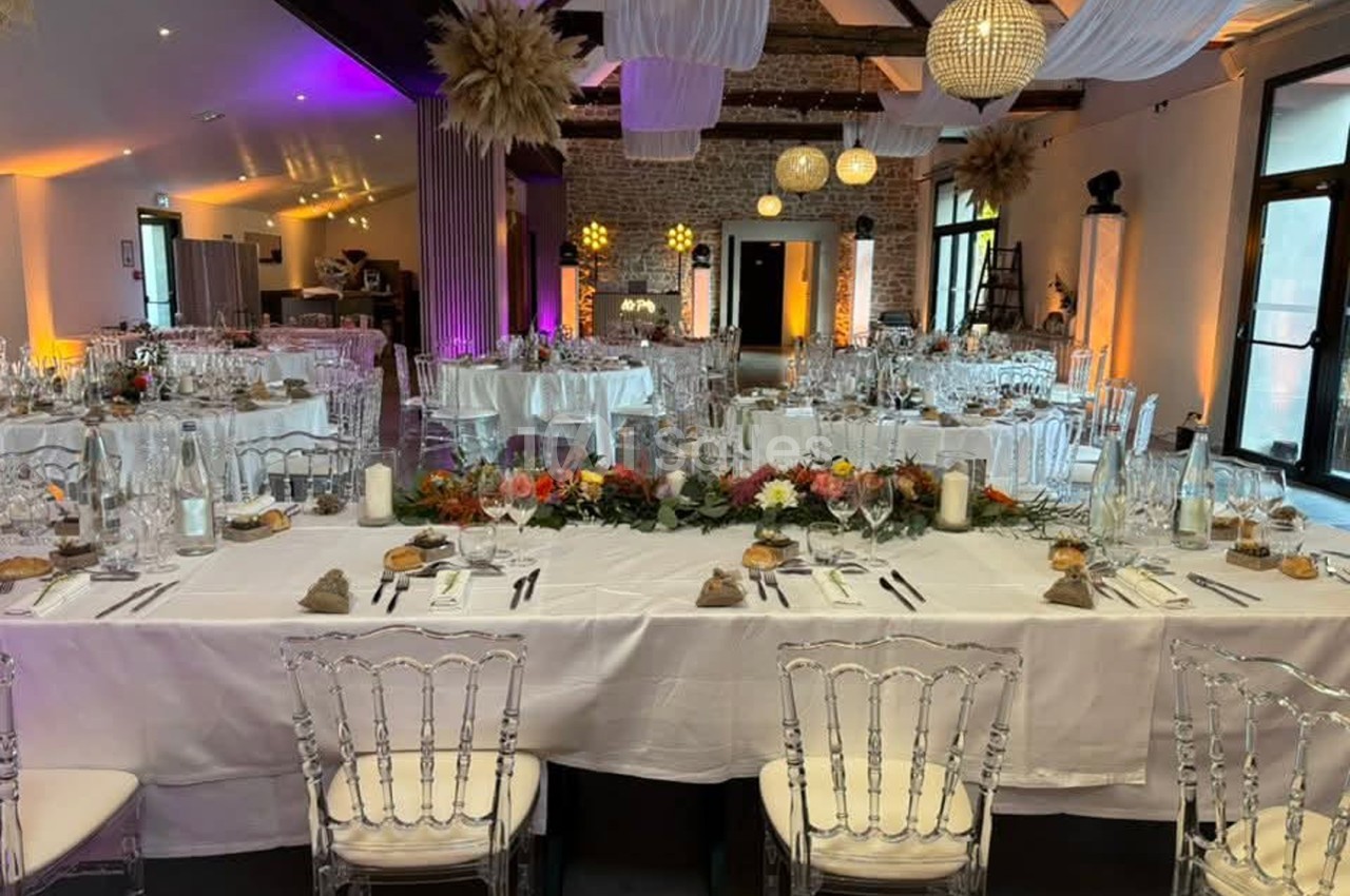 Salle de réception dressée pour un banquet, avec tables rondes et longue table centrale nappées de blanc, chaises transparentes, bouquets colorés et éclairage ambré aux murs de pierre.