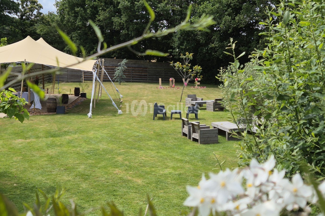 Vaste pelouse en extérieur bordée d'arbres, avec un tipi en toile beige à gauche, un mobilier de jardin en bois et des fauteuils sombres disposés sur l'herbe.