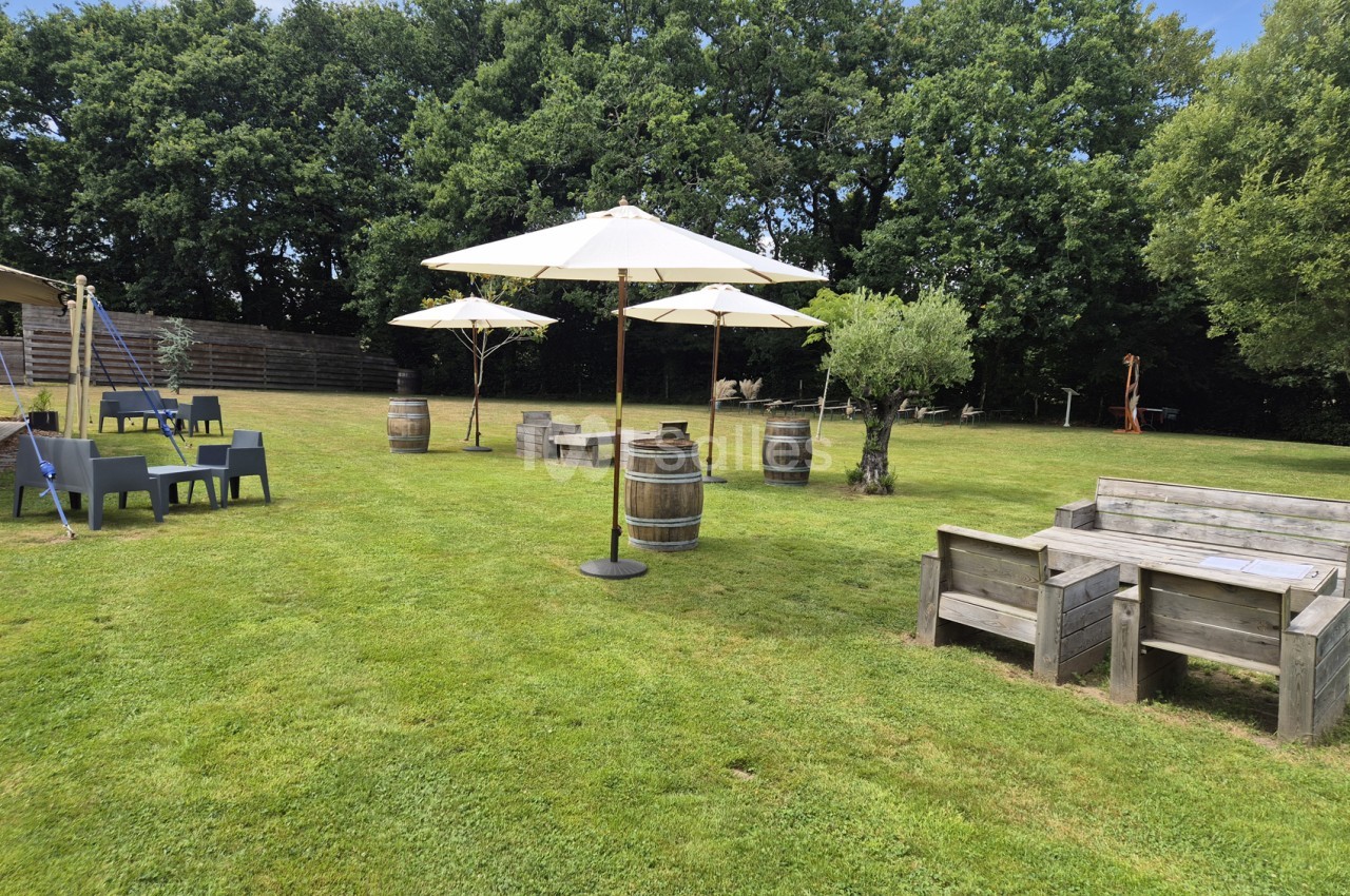 Vaste pelouse d'extérieur ombragée par des grands arbres, avec parasols crème, tonneaux en bois, bancs en bois massifs et quelques fauteuils de jardin en plastique gris.