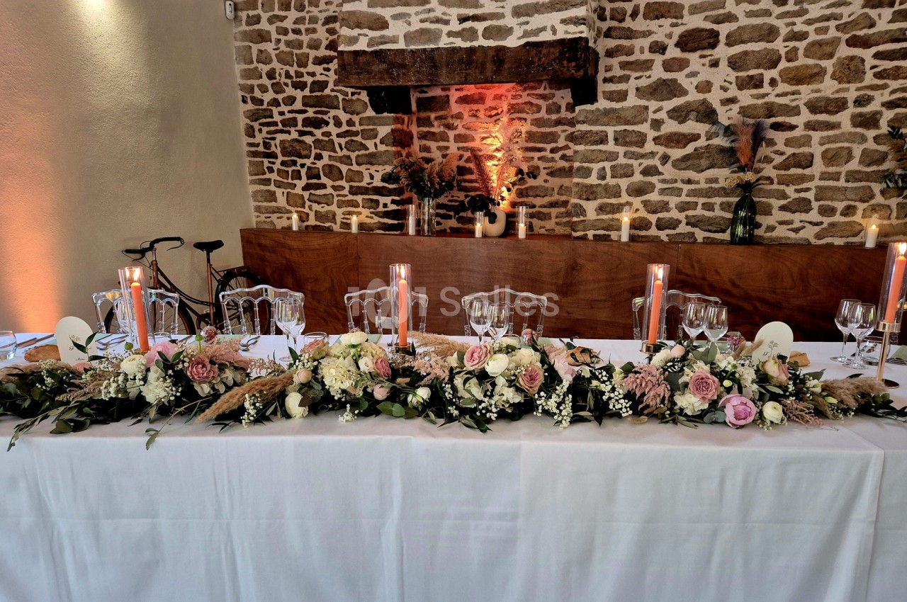 Table d'honneur dressée sous un long centre de fleurs roses et blanches, avec bougies hautes, chaises transparentes et mur de pierres apparentes.