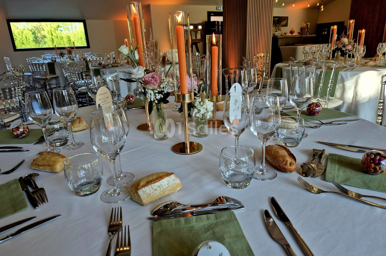 Table ronde dressée de verres à pied, bougies orange sur chandeliers dorés, fleurs roses, serviettes vert sauge et pains posés, dans une salle lumineuse.