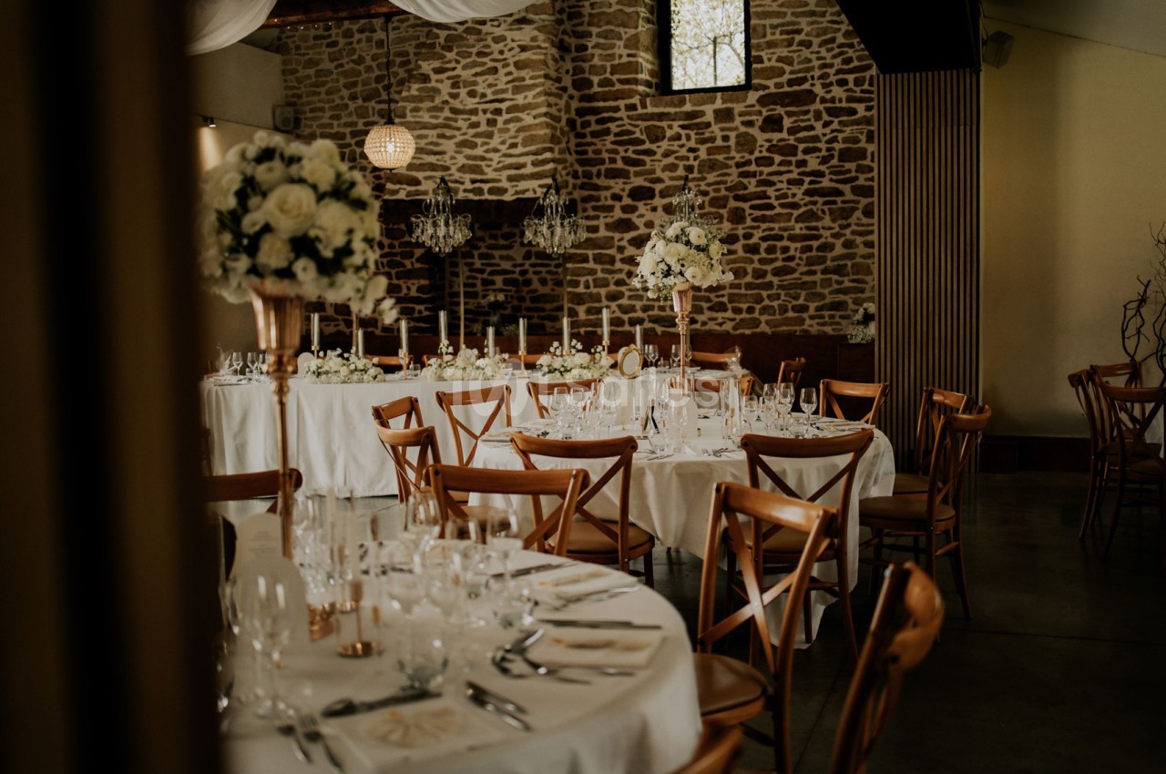 Salle de réception aux murs de pierre apparente, dressée de tables rondes et longues nappées de blanc, avec bouquets de fleurs blanches et chandeliers dorés.