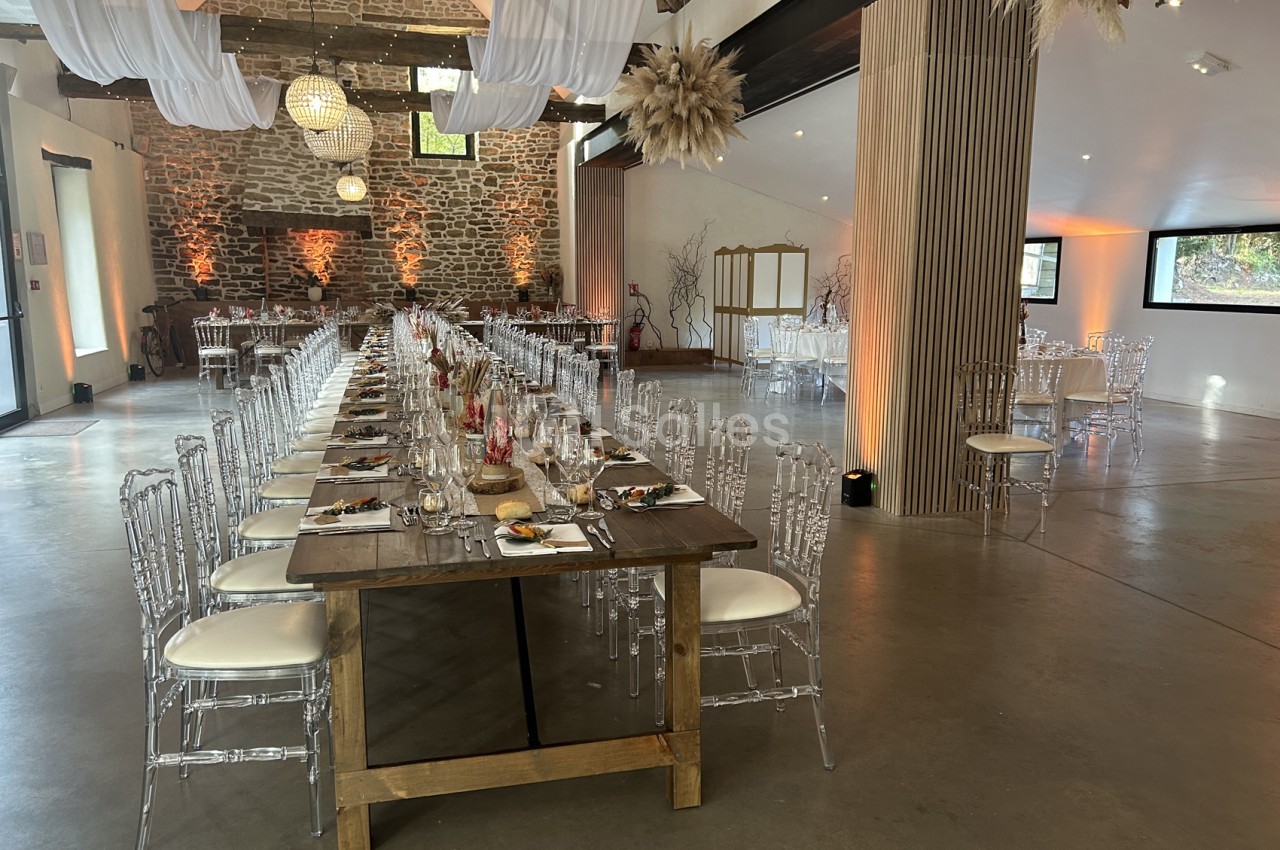 Grande salle de réception avec murs en pierre apparente, longue table en bois dressée de couverts, chaises transparentes à coussins blancs et tables rondes en arrière-plan. Let me verify length: roughly 165 characters, too long. Need ≤125. Salle de