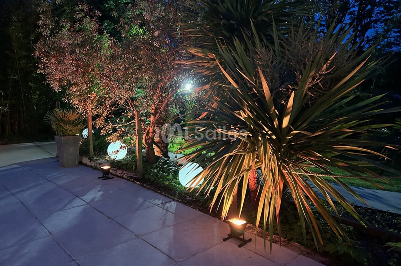 Terrasse carrelée de nuit éclairée par des spots au sol, avec des sphères lumineuses, un palmier yucca et des arbres en arrière-plan.
