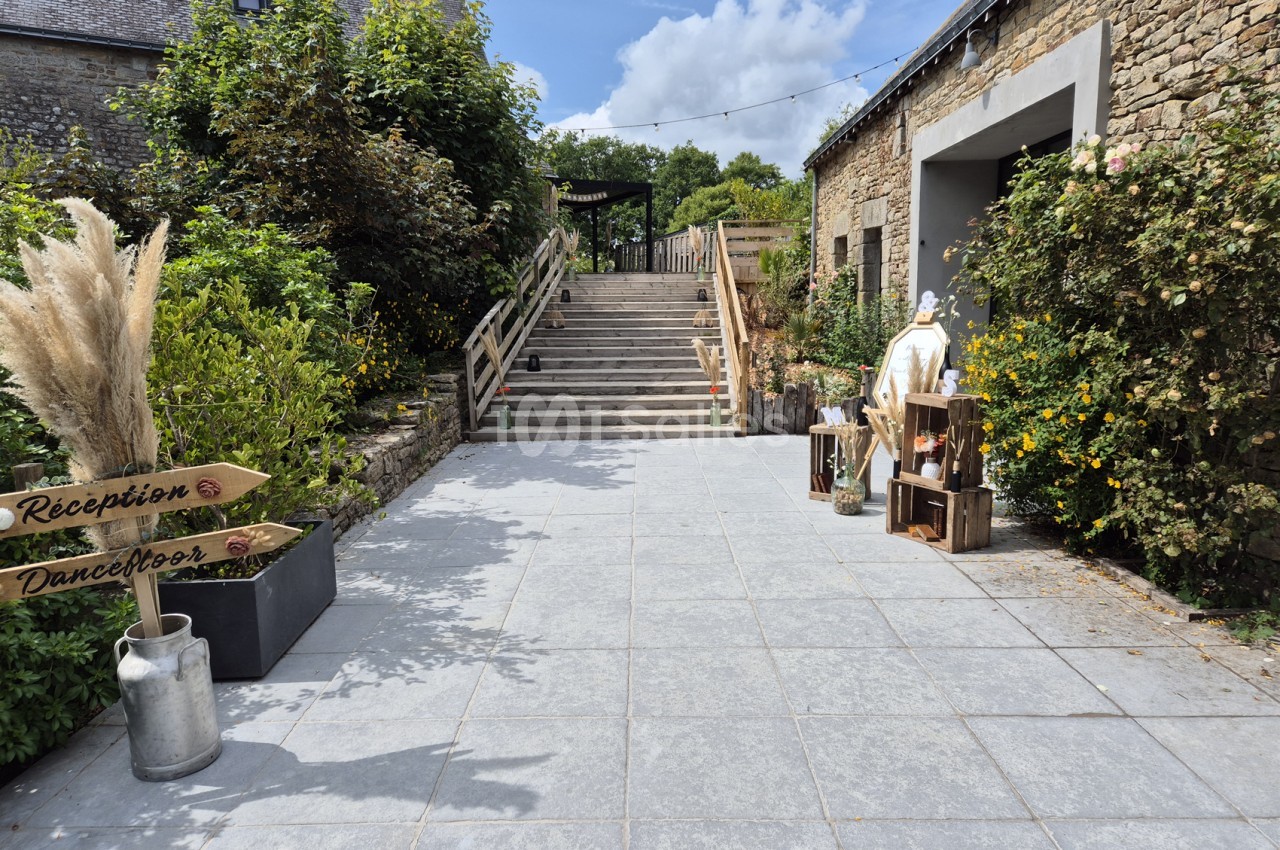 Terrasse pavée menant à un escalier en bois bordé de rambardes, avec des panneaux « Réception » et « Dancefloor » fixés au pot de fleurs près d'un bâtiment en pierre. Wait: I must not transcribe text incrusted in image? The signs are part of the scene