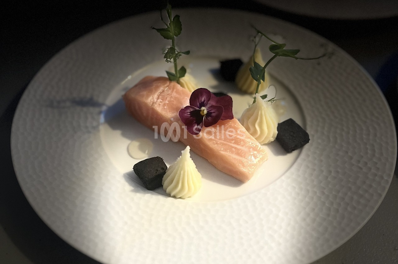 Pavé de saumon rosé dressé sur une assiette blanche texturée, accompagné de quenelles crémeuses, de petits cubes noirs et d'une fleur violette.