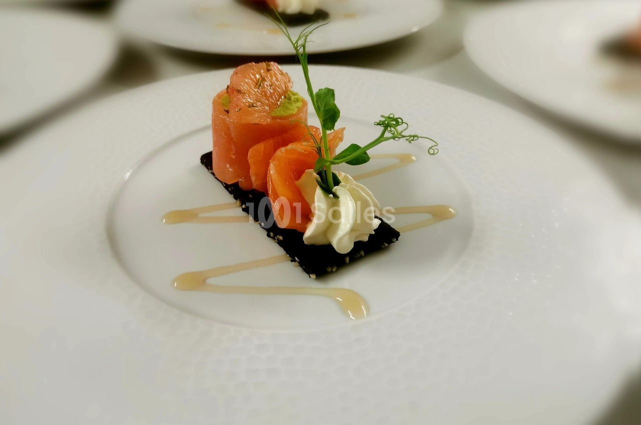 Bouchée de saumon roulé sur un fond noir sésame, accompagnée d'une crème blanche, d'une pousse verte et d'un filet de sauce beige sur assiette blanche.