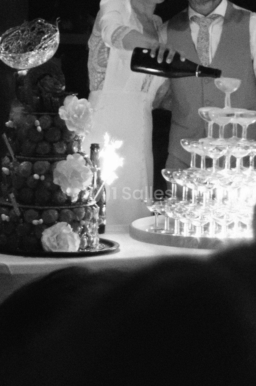 Bouteille de champagne versée dans une pyramide de coupes, près d'une tour de choux ornée de fleurs blanches.