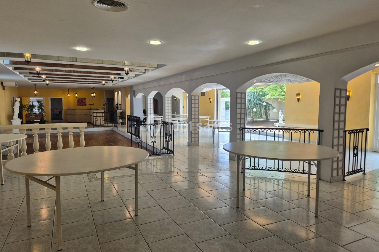 Salle lumineuse avec carrelage clair, tables rondes pliantes et balustrades, donnant sur une terrasse extérieure.