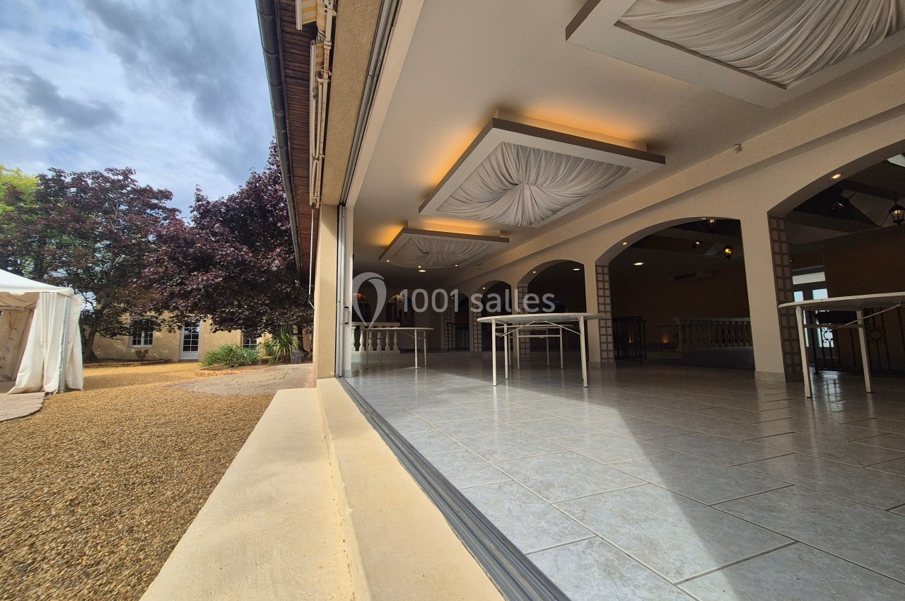 Salle de réception lumineuse avec plafond décoratif, ouverte sur une cour extérieure avec arbres et tente blanche.