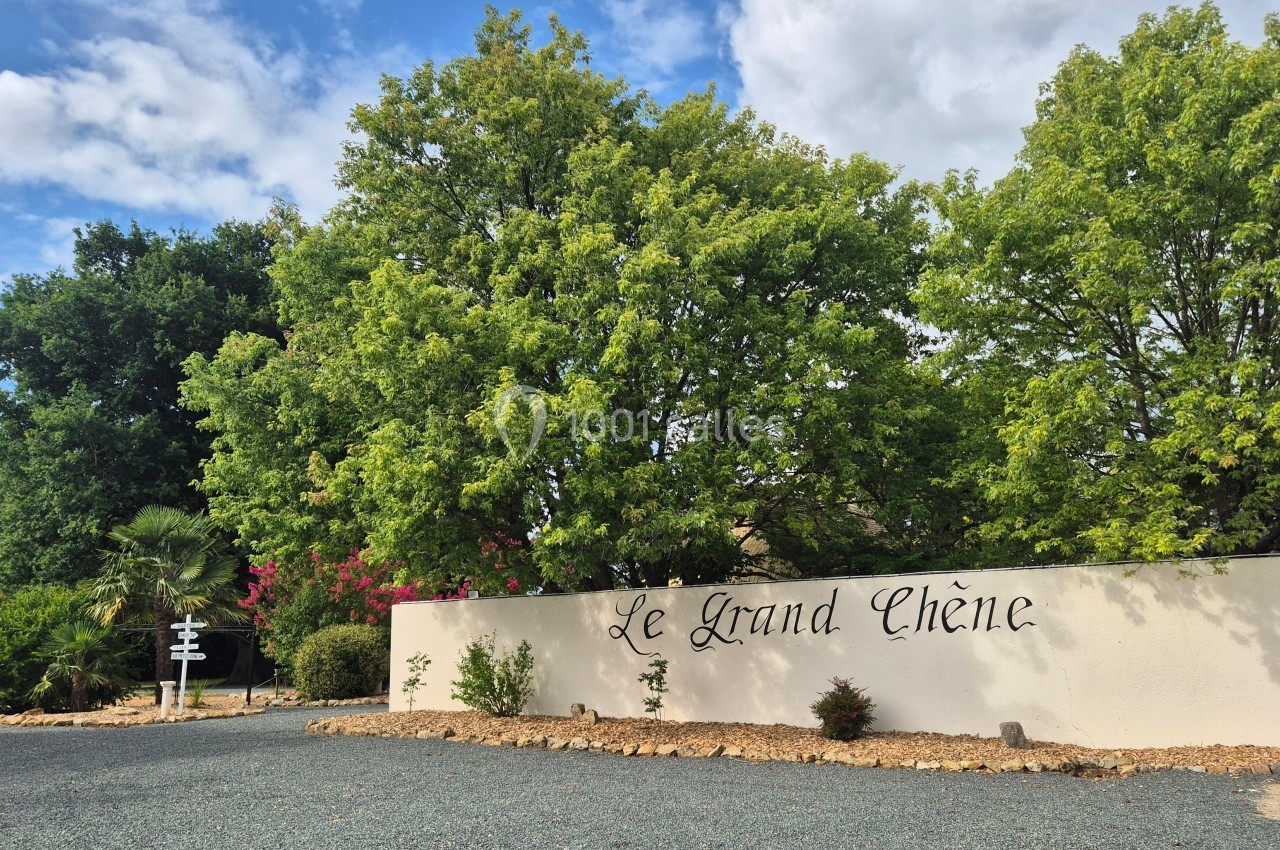 Mur blanc avec l'inscription ’Le Grand Chêne’, entouré d'arbres et d'un chemin gravillonné sous un ciel partiellement…